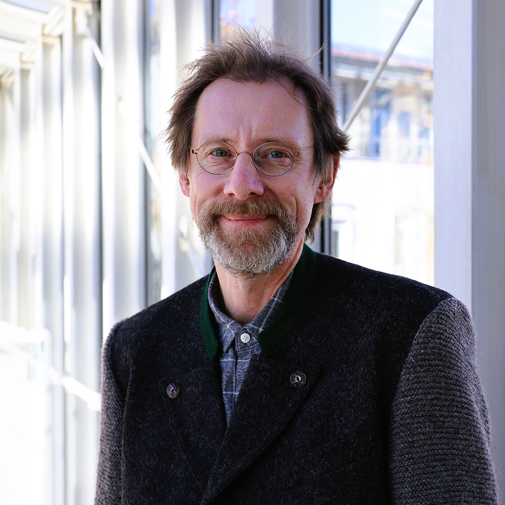 Porträt von Clemens Dannenbeck. Er trägt eine Brille, dunkelbraunes Haar und einen grau-braunen Bart. Außerdem ein Hemd und eine legere Jacke darüber.