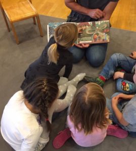 Eine Person kniet am Boden und hält das aufgeschlagene Kinderbucht. Rund herum sitzen fünf Kinder. Ein Mädchen beugt sich nach vorne und zeigt auf das Buch.