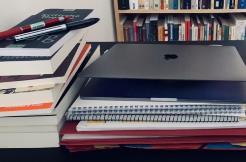 Im Hintergrund sieht man ein Bücherregal. Davor einen Stabelbücher. Darauf liegen zwei Stifte. Neben dem Bücherstapel liegt ein Stapel mit Maßen und Hefte. Darauf liegt ein leicht aufgeklapptes Laptop.