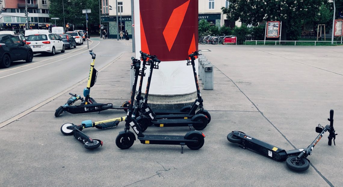 Mehrere E-Scooter liegen am Gehsteig herum oder lehnen an einer Litfaßsäule