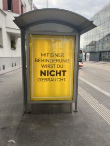 Man sieht eine überdachte Bushaltestelle von der Seite. Es ist ein Plakat in einer Werbefläche angebracht. Es ist leuchtend gelb. Darauf steht mit schwarzer Schrift: Mit einer Behinderung wirst du nicht gebraucht.