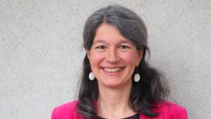 Ein Porträt von Susanne Jaquemar. Sie trägt ein Pinkes Oberteil und Ohrringe. Graue schulterlange Haare. Sie lächelt in die Kamera.