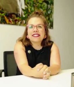 Portraitfoto von Ninia Binias. Sie trägt eine Brille und knalligen Lippenstift. Sie hat ihre Arme auf dem Tisch abgestützt und lächelt in die Kamera.