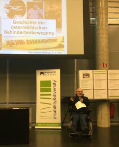 Volker Schönwiese spricht zum Publikum. Hinter ihm sieht man seine Powerpoint-Präsentation und ein Roll-Up des disability&diversity Zentrums Salzburg.